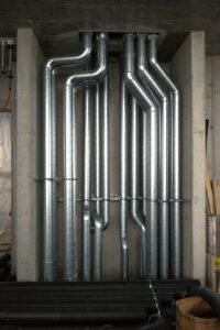 Technicians installing large ductwork and piping inside a spacious industrial facility.