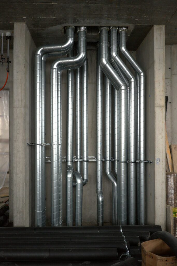 Technicians installing large ductwork and piping inside a spacious industrial facility.