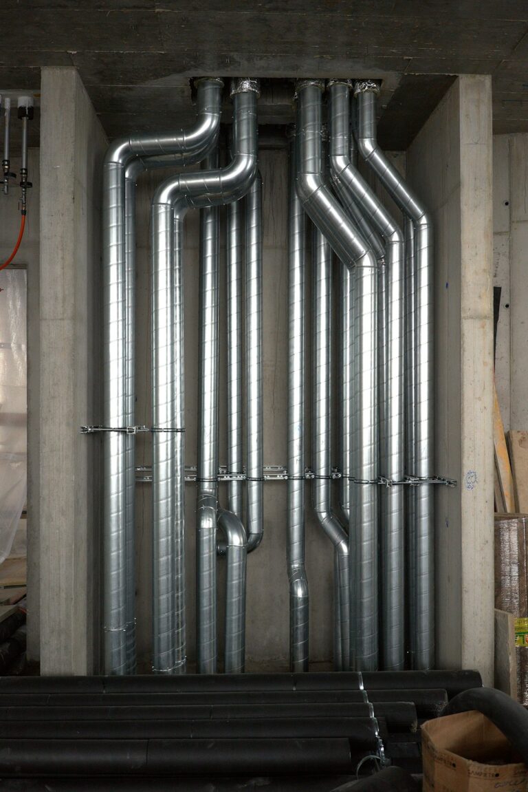 Technicians installing large ductwork and piping inside a spacious industrial facility.