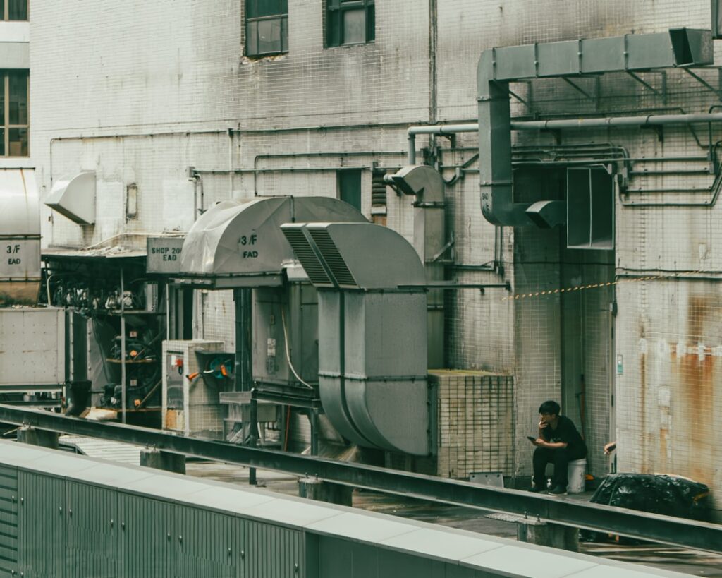 A construction worker reviews mechanical blueprints next to exposed pipes and ductwork in a building under renovation.