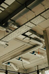 Engineers review mechanical drawings beside ductwork, pipes, valves, and heating equipment in a building work area.