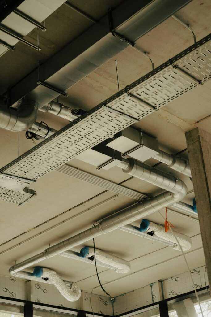 Engineers review mechanical drawings beside ductwork, pipes, valves, and heating equipment in a building work area.