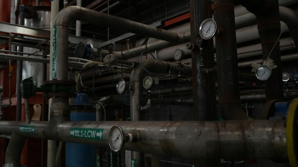 Workers assemble labeled pipe sections and duct components on a fabrication-shop floor beside tools and metal supports.