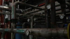 Workers assemble labeled pipe sections and duct components on a fabrication-shop floor beside tools and metal supports.